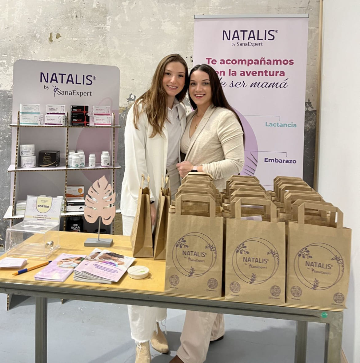 Laura und Nayara von SanaExpert lächeln hinter einem Natalis-Produktstand mit einer Auswahl an Gesundheitsprodukten und informativen Broschüren, umgeben von Papiertüten.