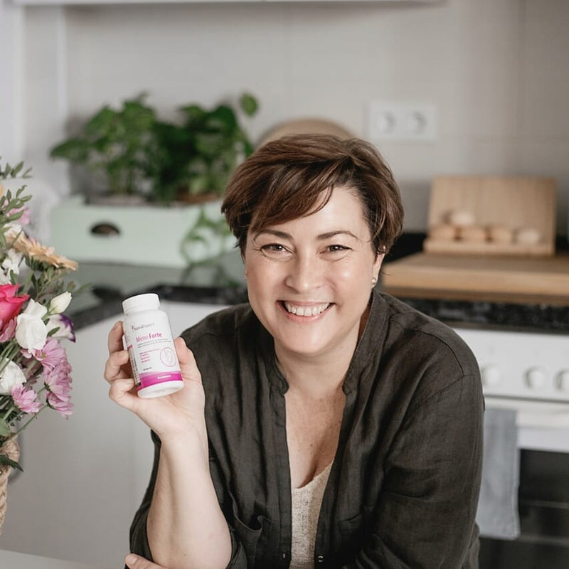 Eine Frau mittleren Alters hält ein Produkt von SanaExpert Meno Forte in der Küche, umgeben von Blumen und Küchenutensilien, freundliche und heitere Atmosphäre.