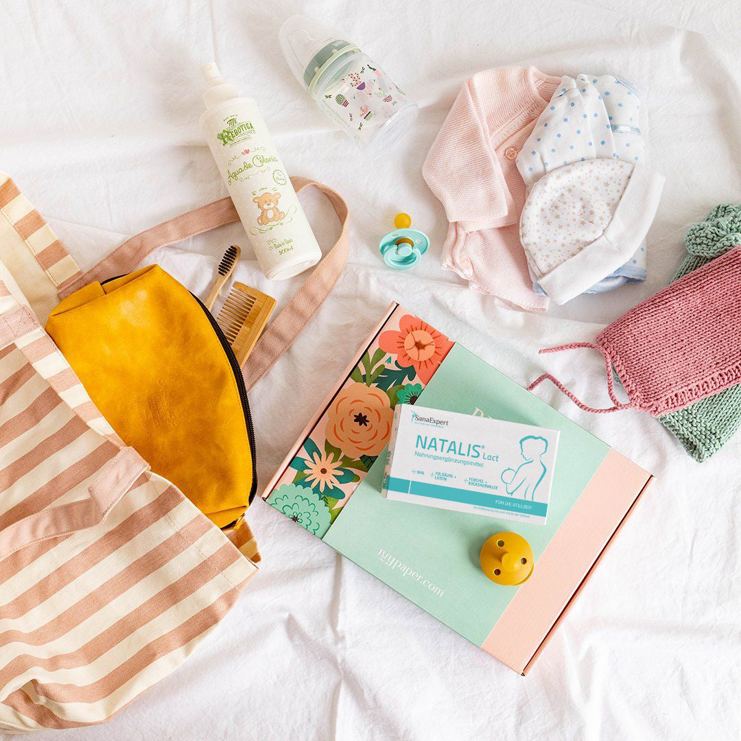 Wickeltasche, Babypflegeprodukte, Schnuller und eine Packung Natalis Lact auf einem Bett mit weißem Hintergrund.