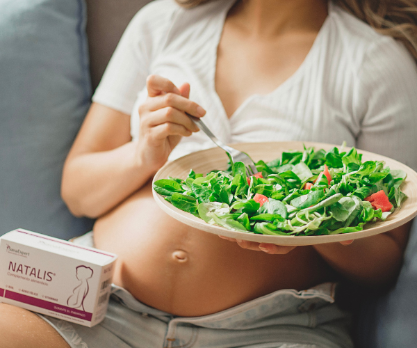 Schwangere Frau sitzt auf einem Sofa und isst Salat, während eine Packung Natalis neben ihr liegt.