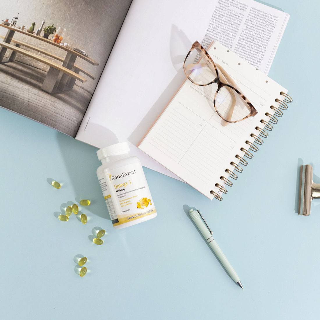 SanaExpert Omega-3 Kapseln verstreut neben einem offenen Notizbuch und einer Brille auf einem Schreibtisch, symbolisiert die Verbindung zwischen Gesundheit und produktiver Arbeit.