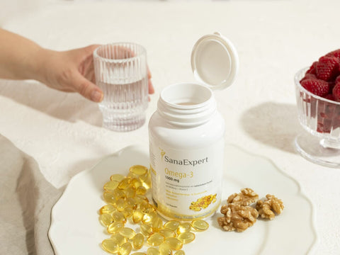 Eine geöffnete Flasche SanaExpert Omega-3 neben einem Teller mit goldenen Kapseln, eine Hand hält ein Glas Wasser, Himbeeren und Walnüsse ergänzen das Bild für eine gesunde Ernährung.