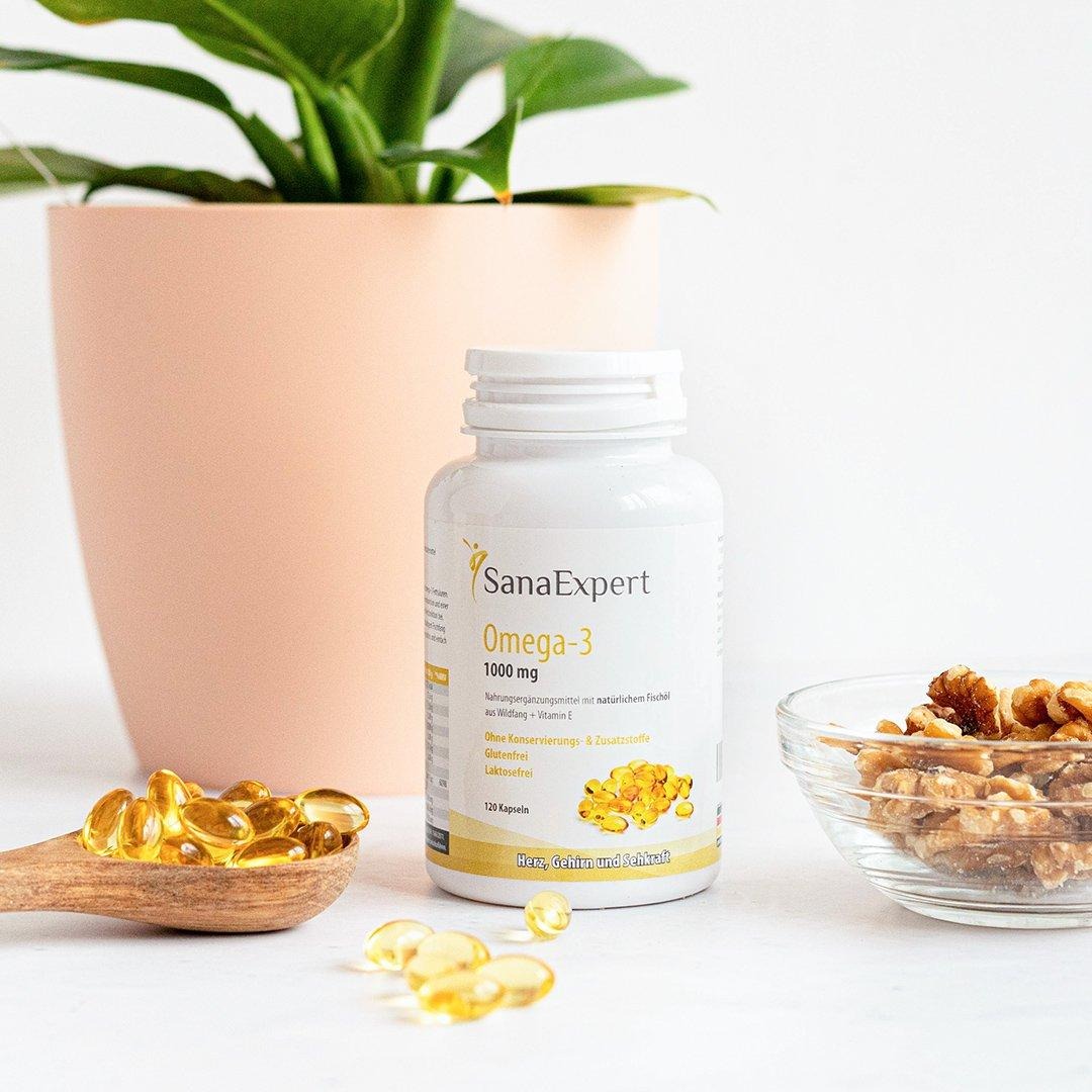 SanaExpert Omega-3 Flasche neben einer hölzernen Schale mit goldenen Fischölkapseln und einer Schüssel mit Walnüssen, vor einer großen Topfpflanze, die Natürlichkeit unterstreicht.