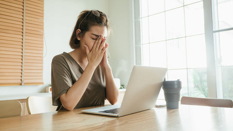 Junge Frau sitzt erschöpft am Schreibtisch vor ihrem Laptop und reibt sich die Augen, ein Bild, das digitale Überanstrengung und die Notwendigkeit von Pausen veranschaulicht.