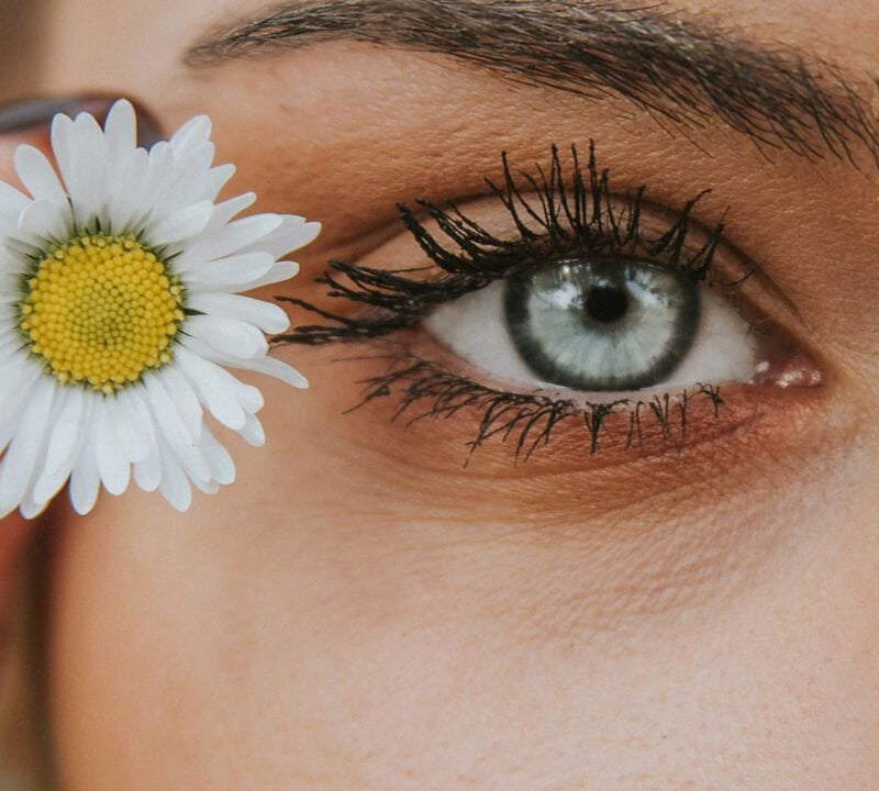 Detailaufnahme eines weiblichen Auges mit einer Gänseblümchenblüte, unterstreicht visuelle Gesundheit und Natürlichkeit.