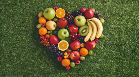 Ein herzförmig angeordnetes Arrangement aus verschiedenen Früchten, darunter Äpfel, Orangen, Bananen, Trauben und Beeren, auf einem grünen Rasenhintergrund.