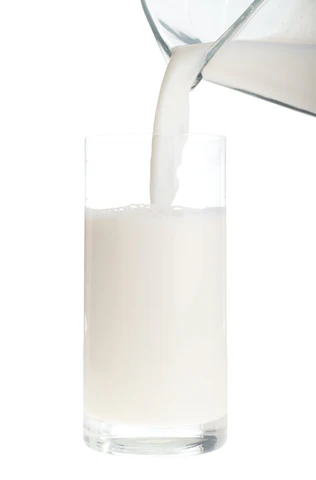 Milch wird aus einem Edelstahlkrug in ein klares Glas gegossen. Der weiße Milchstrahl bildet einen Kontrast zum transparenten Glas und dem hellen Hintergrund.