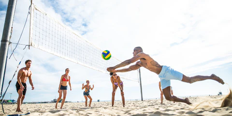 Eine dynamische Gruppe von sechs jungen Erwachsenen spielt Beachvolleyball am sonnigen Strand, wobei ein Spieler spektakulär nach dem Ball hechtet.