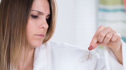 Eine junge Frau mit langen, glatten Haaren zeigt besorgt auf eine Ansammlung von Haaren auf ihrer Schulter, ein Anzeichen für Haarausfall.