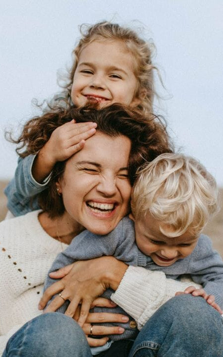 Mutter lacht und umarmt ihre zwei Kinder liebevoll am Strand, ausgelassene und freudige Familie.
