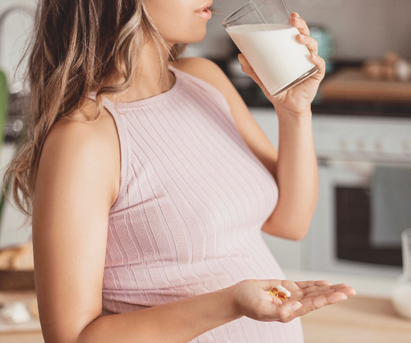 Schwangere Frau in einem rosa Kleid steht in der Küche und hält ein Glas Milch in einer Hand und Nahrungsergänzungsmittel in der anderen.