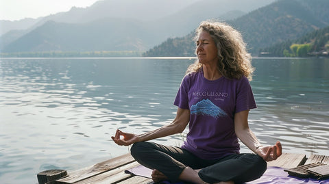 Eine Frau in einem lila T-Shirt meditiert in einer sitzenden Pose auf einem Holzsteg am See, umgeben von ruhigem Wasser und grünen Bergen.