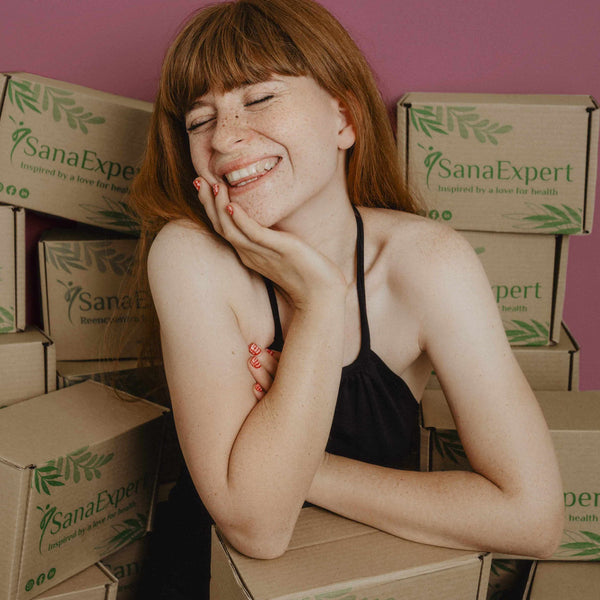  Eine junge Frau mit sommersprossen und roten Haaren lehnt lachend an einem Stapel von SanaExpert Produktkartons.