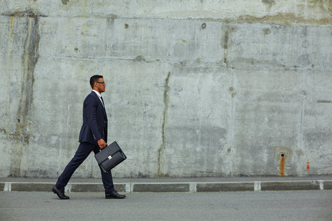Ein Geschäftsmann in einem dunkelblauen Anzug und mit einer Aktentasche geht entschlossen an einer großen grauen Betonwand entlang. Er trägt eine Brille und schaut konzentriert nach vorne, während er auf einer leeren Straße läuft.