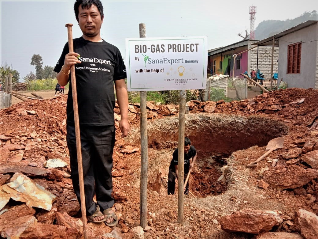 Zwei Männer bei der Arbeit an einem Biogas-Projekt, neben einem Schild mit der Aufschrift "BIO-GAS PROJECT by SanaExpert Germany with the help of", vor einer großen Grube im Erdboden. Der vordere Mann hält eine Schaufel und trägt ein T-Shirt mit dem SanaExpert-Logo.