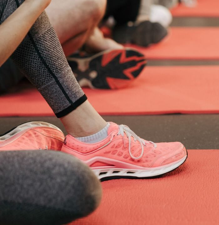 Teil eines Fitnessstudios mit Fokus auf die Beine einer Person, die auf einer roten Gymnastikmatte sitzt, neben ihr ein Paar rosa Sportschuhe.