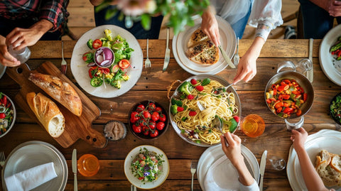 Eine Gruppe von Menschen genießt gemeinsam eine Mahlzeit an einem rustikalen Holztisch, bestückt mit einer Vielzahl von gesunden Gerichten wie Salaten, Nudeln und frischem Gemüse.