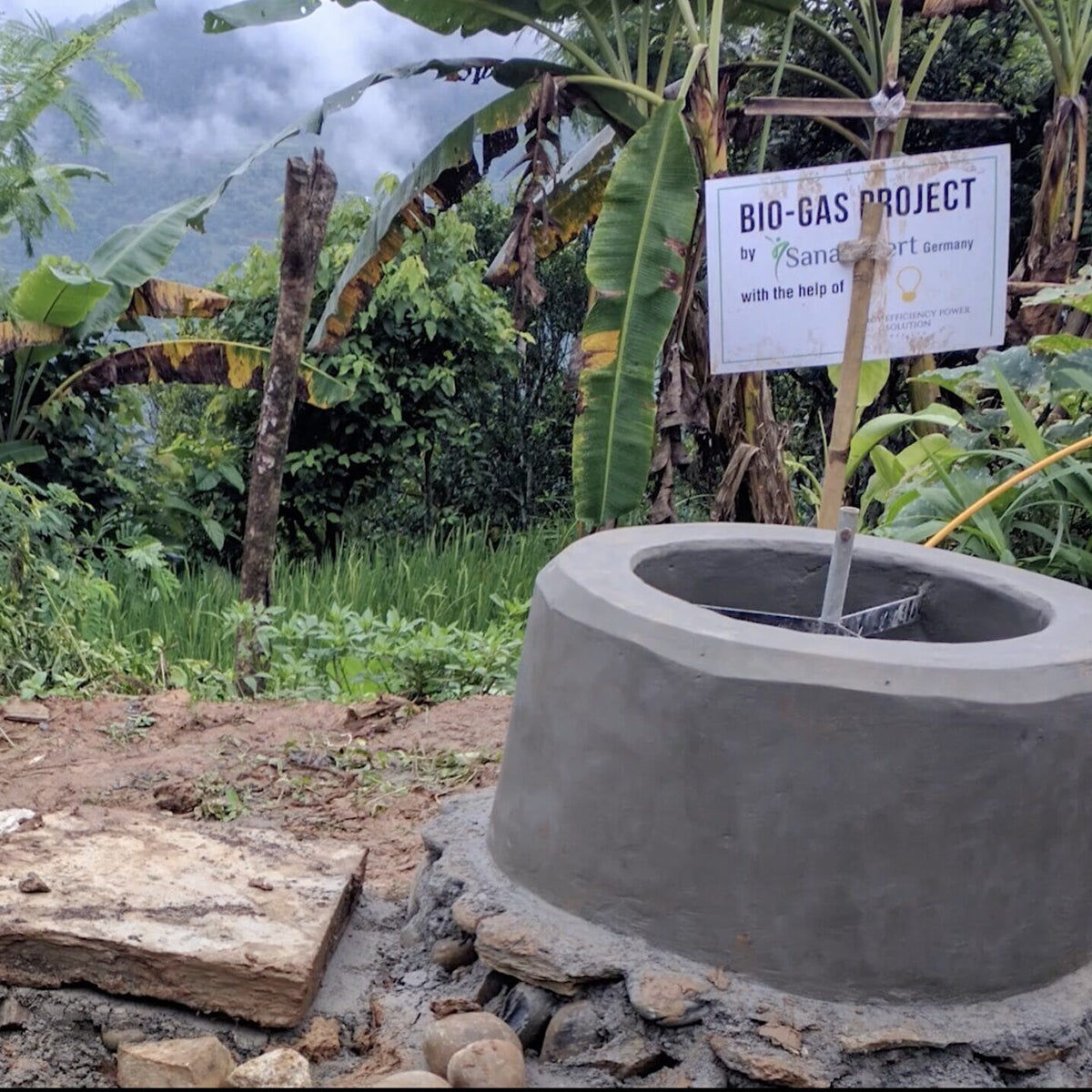 Biogas-Anlage im Freien mit einem Schild, das das Biogas-Projekt von SanaExpert ankündigt, umgeben von tropischer Vegetation.