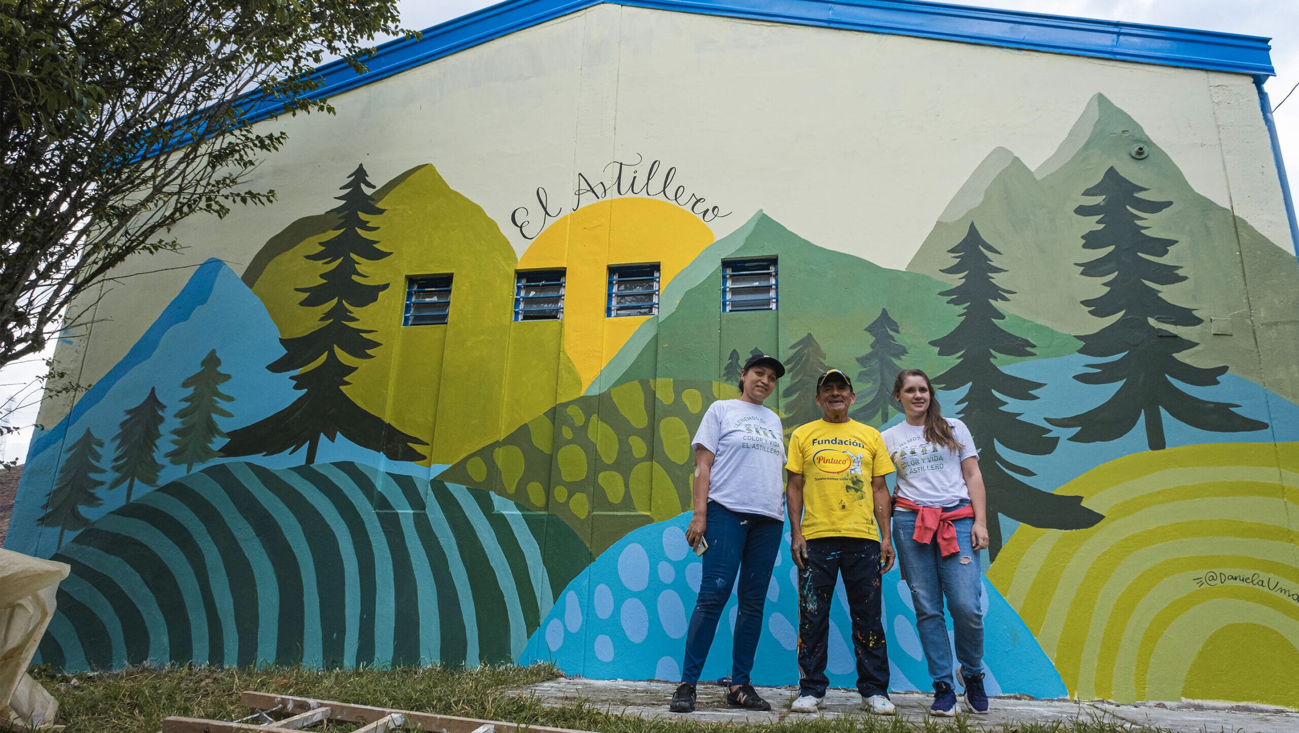 Drei Freiwillige stehen stolz vor einer großen Wandmalerei mit bunten, abstrakten Bergen und Bäumen, der Schriftzug "El Astillero" ist über ihnen an der Wand zu sehen.