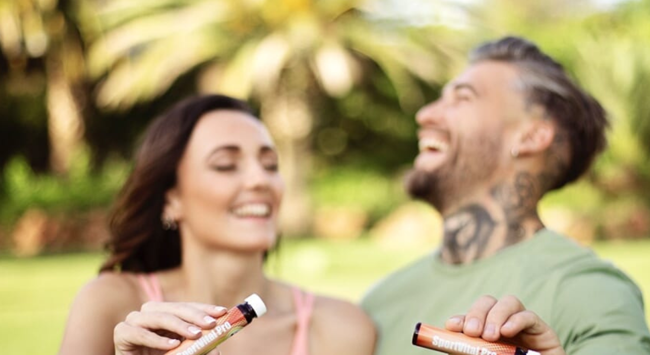 Lächelnde Frau und Mann im Freien halten SportVital Pro Nahrungsergänzungsmittelröhrchen, unscharfer grüner Hintergrund.