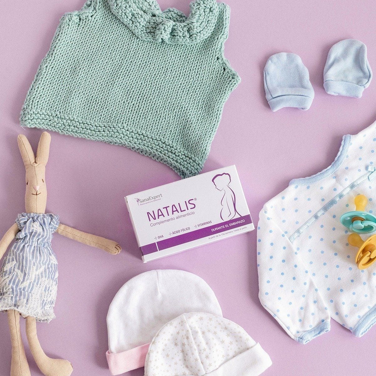 Flachgelegte Anordnung von Babykleidung und Natalis Nahrungsergänzungsmitteln auf einem rosa Hintergrund, bereit für die Ankunft eines Neugeborenen.