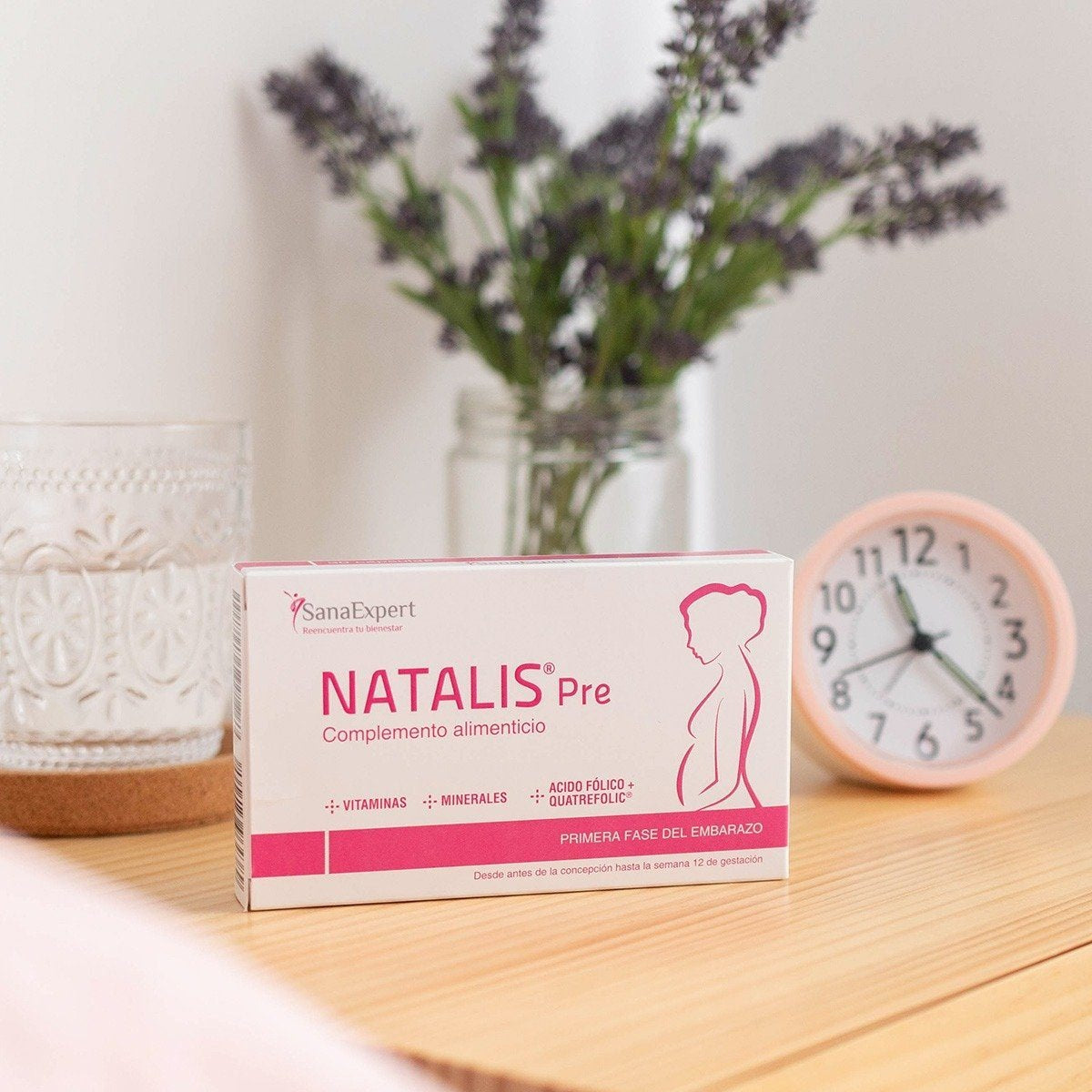 Verpackung von Natalis Pre Nahrungsergänzungsmitteln für Schwangere auf einem Tisch neben einer Uhr und einer Vase mit Lavendel.