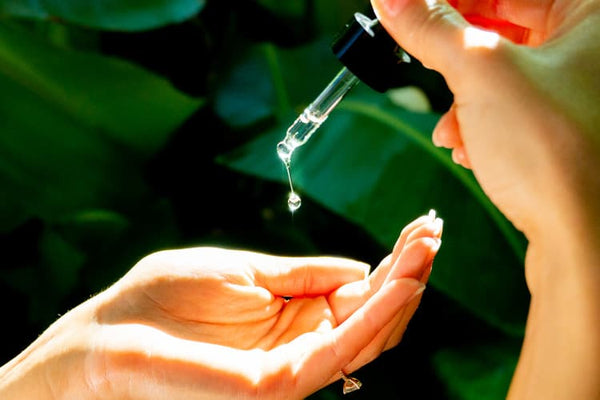 Hand using a face oil filled dropper onto other palm with green plants in the background
