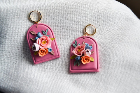 a pair of arch-shaped magenta clay earrings with peach, pink, orange, and lavendar florals on a bright white fabric backdrop