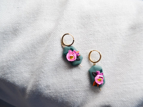 a pair of elongated hexagonal clay earrings in a medium teal color with orange, black, and dark green leaves and hot pink and purple flowers. Earrings are sitting on a bright white fabric background