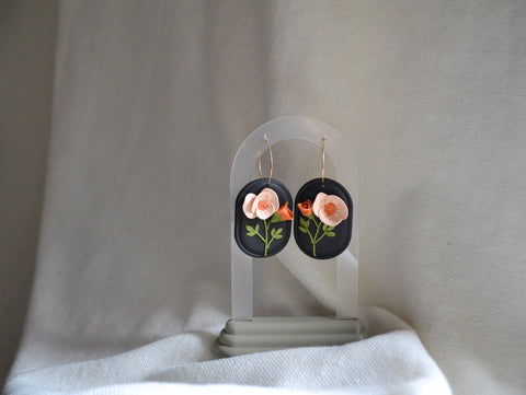 a pair of clay earrings with a black background and peachy poppies hangs on a translucent arched earrings holder
