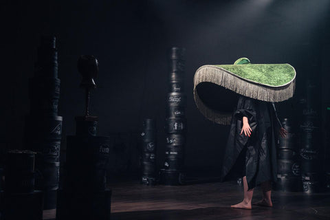 A performer in a black poncho stands in a set of hat boxes wearing a large green hat with fringe trimming as part of the production of Far Away by Patalog Theatre