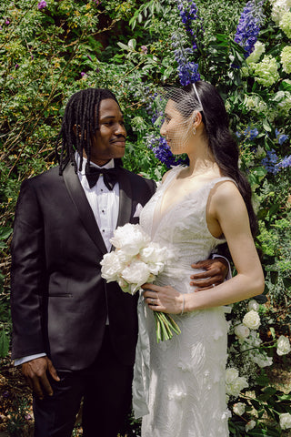 A bride and groom stand in a lush green garden the bride is wearing a face veil