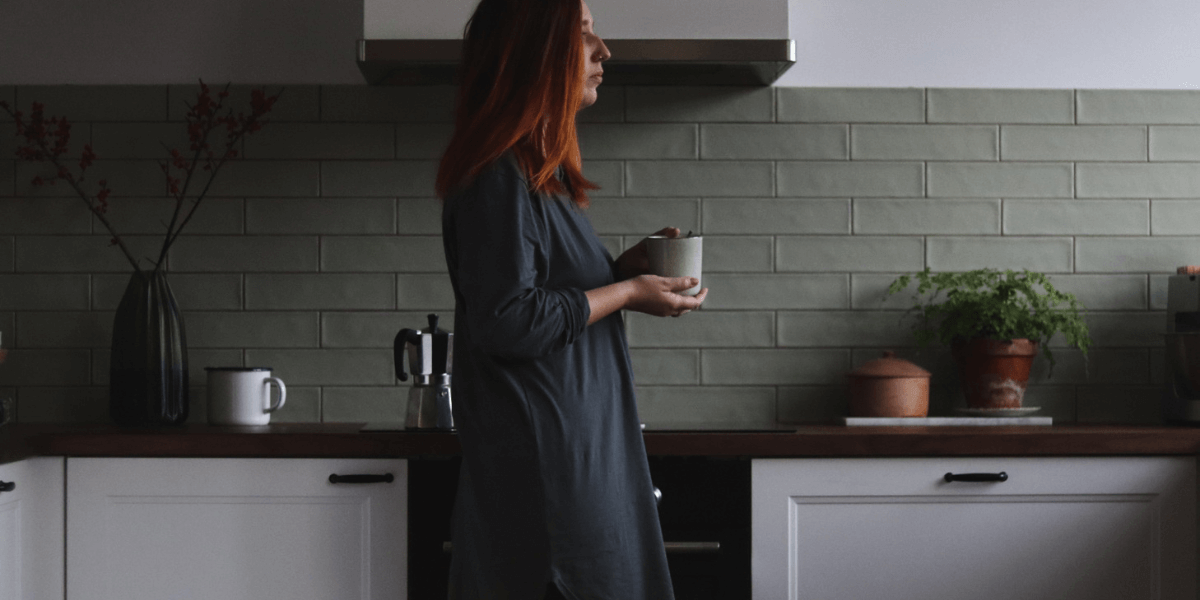 Kim standing in their kitchen in a long grey shirt (which is actually a nightshirt but don't tell anyone), holding a cup of coffee, staring out the window.