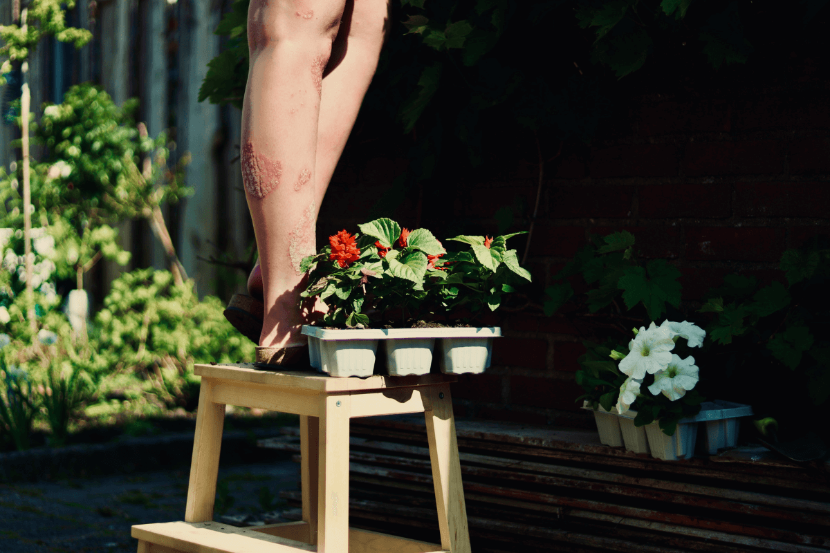 Kim's lower legs on a wooden step stool in the garden, a tray of red and white petunias next to their feet. They are planting these plants out of frame.