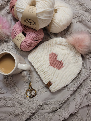 A photo showing a white beanie with a pink heart detail and pink pompom on it. Next to the beanie is a cup of tea and 3 balls of Stitch & Story The Chunky Wool.