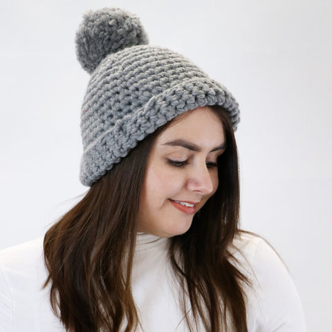 Image shows a woman wearing a grey crochet beanie with a pom pom on top.