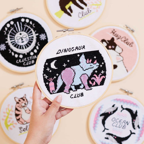 Image shows 6 cross stitch hoops with find your club designs in them. Holding closest to the camera is a Dinosaur club cross stitch design using black, light blue, lilac and fuchsia pink as accent.