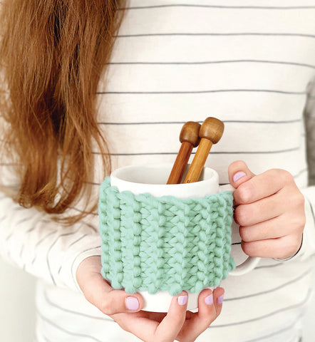 Woman holding a mug with a pair of knitting needles in it, and a Stitch & Story knitted cup cosy around the mug.