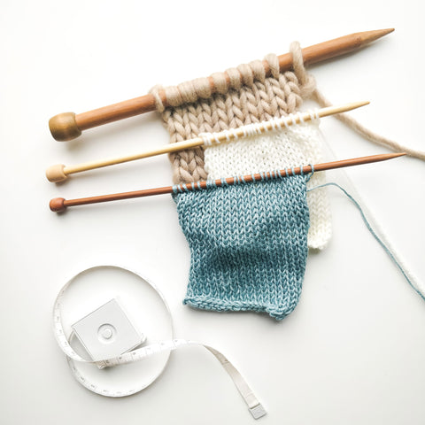 Image showing 3 knitted swatches still on needles, all in stocking stitch, and a measuring taped styled at the bottom left of the image