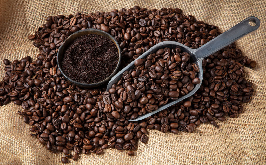 Bowl of coffee grounds next to pile of whole coffee beans laying on burlap.