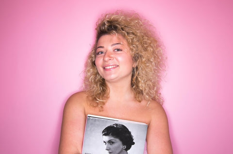 mulher loira sorrindo e segurando uma revista em frente a uma parede rosa