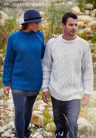 Image of cover of Sirdar knitting pattern 9465 featuring a man and a woman wearing hand knitted Aran sweaters. One in a dark blue and the other in a natural shade