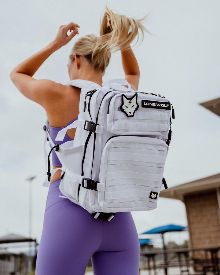 Blonde woman fixing her hair wearing a purple cropped top, leggings, and a white athletic backpack 