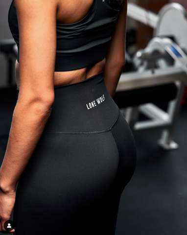Woman wearing black athletic leggings with Lone Wolf written in white text across the back and a black sports bra at the gym