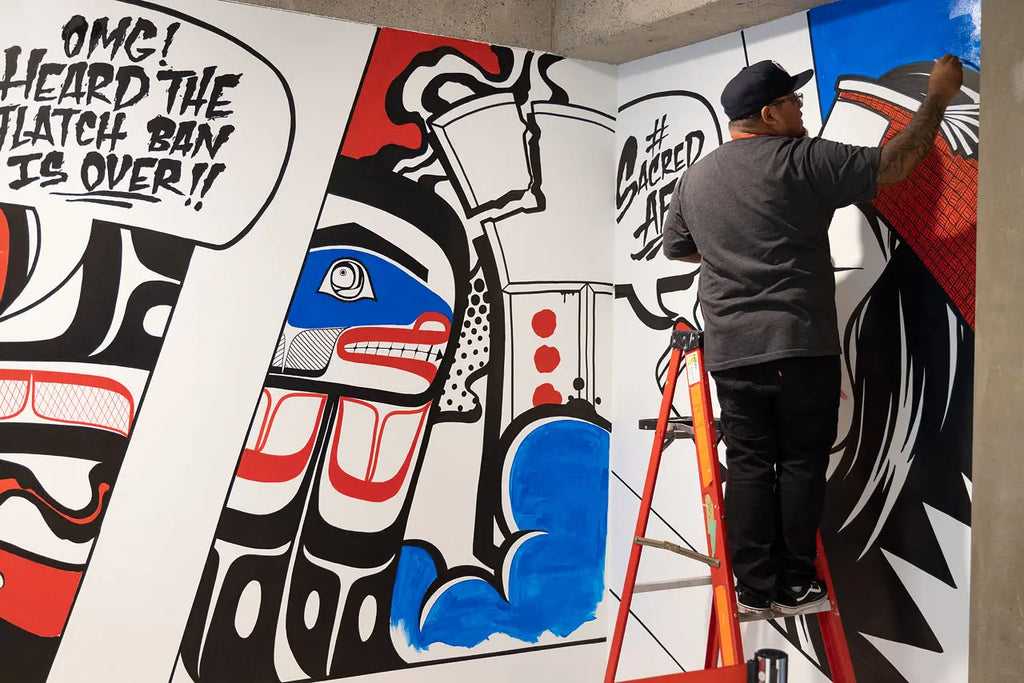 [Image Description: KC Hall stands on a ladder, adding finishing touches to his mural in the Gallery Lobby. The mural is painted across 2 perpendicular walls in a Pop Art and graffiti style, infused with Pacific Northwest Coast art elements. The colours red, black, white and blue are dominant. In the top left corner, a word bubble contains a statement that reads: “Omg! I heard the potlatch ban is over!” On the right, another word bubble contains the statement: “#Sacred A F.”]