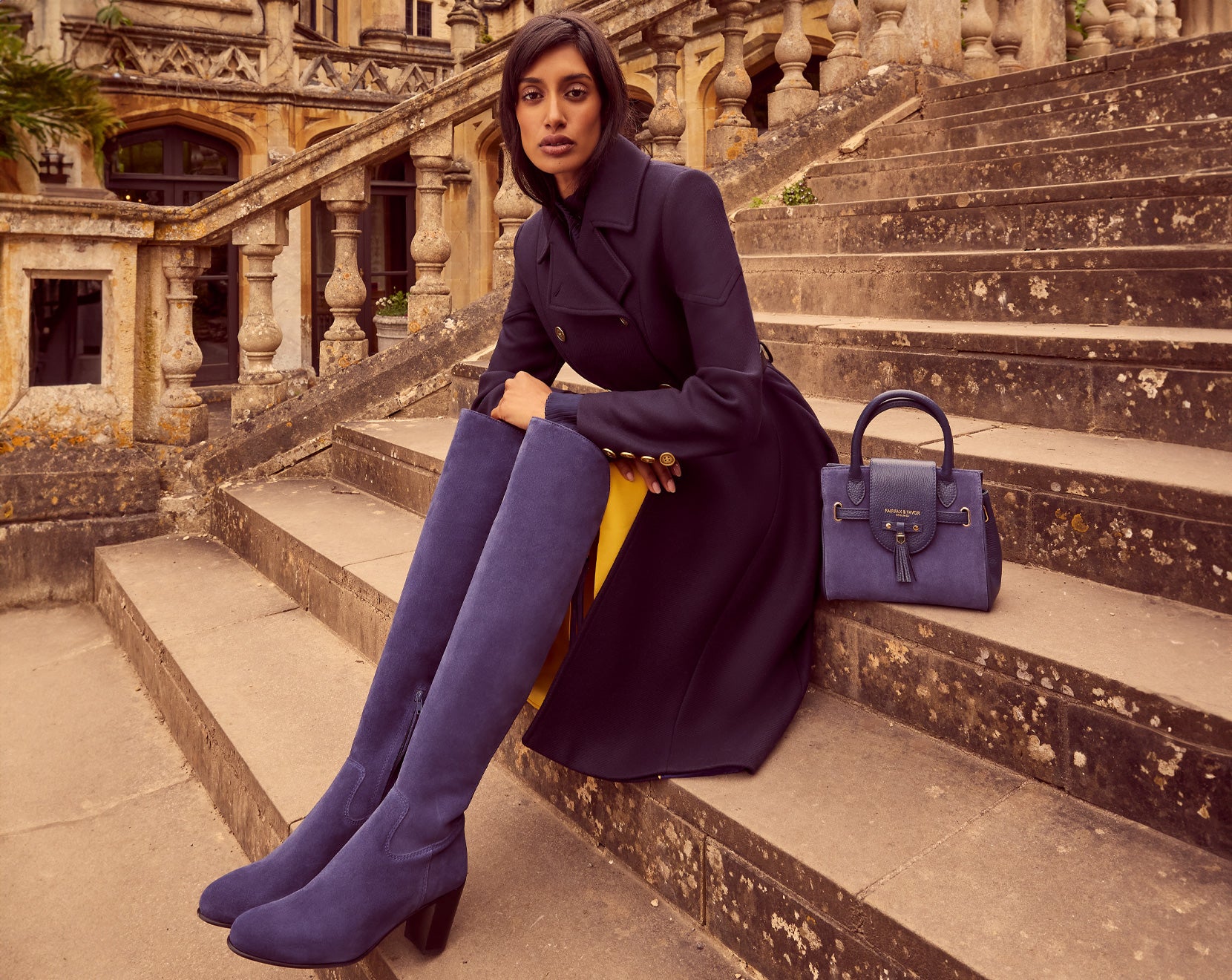 The model is wearing the Victoria coat in navy wool, the Mini Windsor crossbody handbag in ink suede and the Heeled Amira Boots in ink suede looking classy.