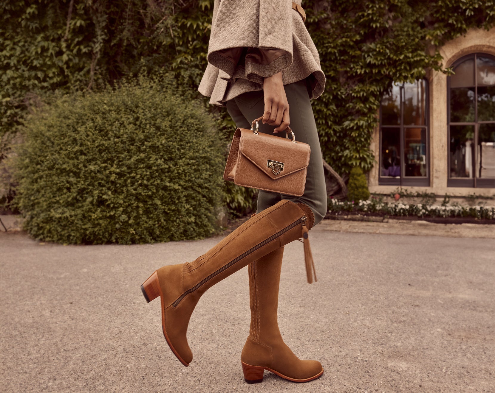 The Model is wearing the Sienna cape in taupe, the Mini loxley in tan leather and the heeled Regina boot in tan suede, looking refined and elegant.