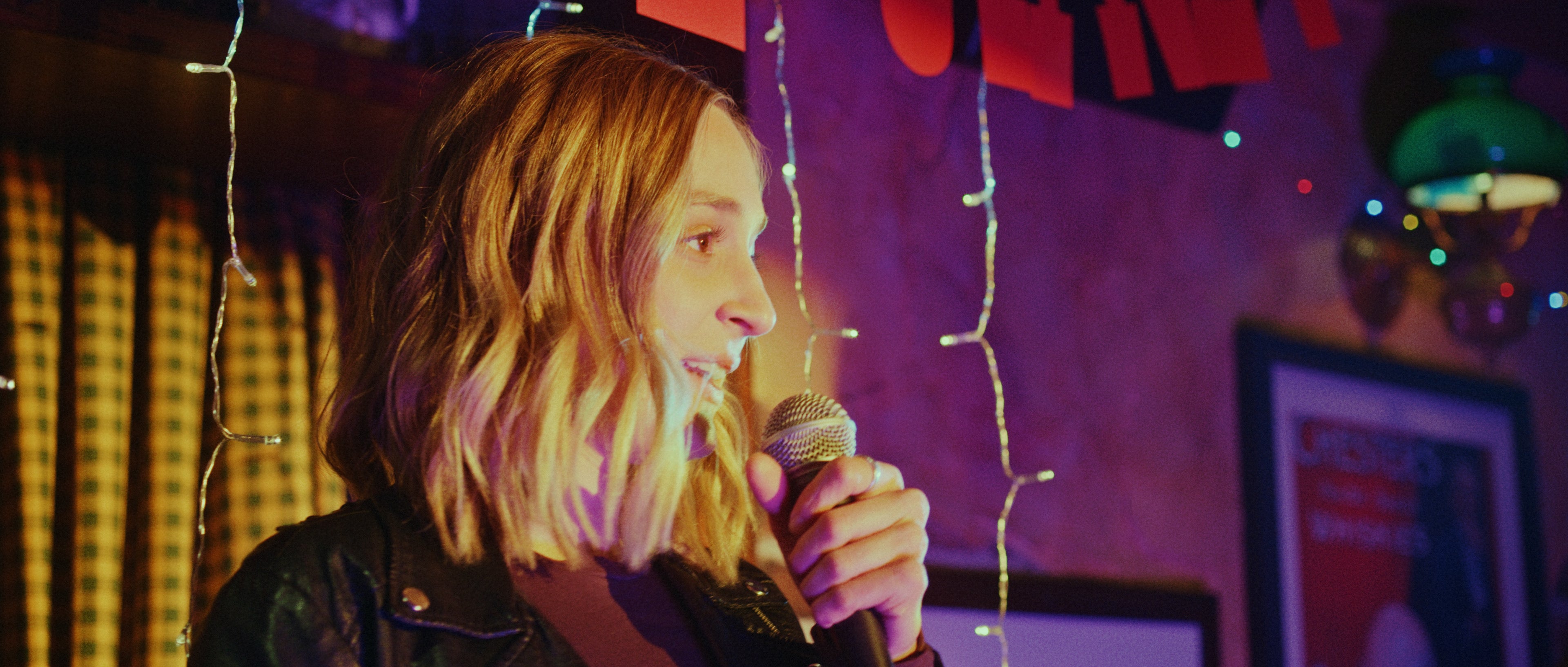 A still from Will Schneider's Goodbye Janet - a young woman grips a microphone while in the spotlight of a karaoke bar