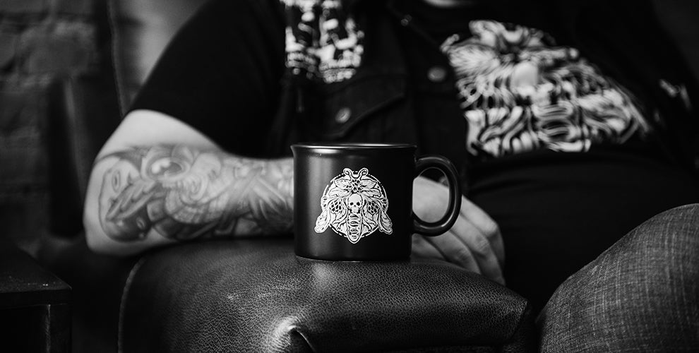 A tattoo artist with a sleeve of tattoos holding a Death Wish Coffee mug with his tattoo design on the front.
