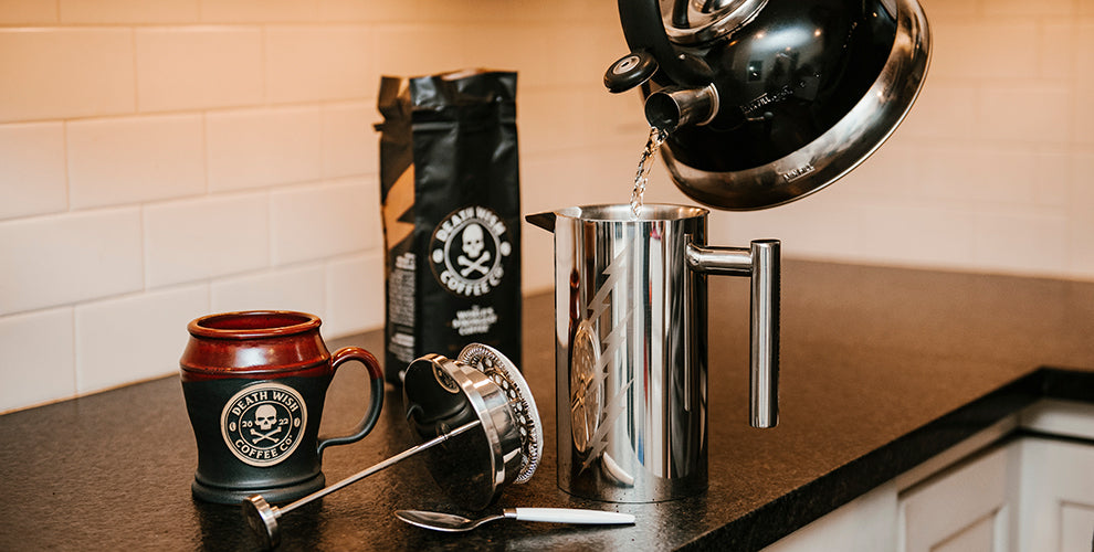 A kettle being poured into a silver French press next to a bag of Death Wish Coffee.